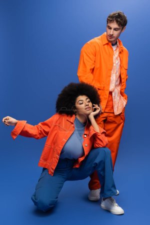 Stylish african american woman posing near man in jacket and overall on blue background