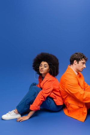 Foto de Curly african american woman in bright orange jacket sitting near man on blue background - Imagen libre de derechos
