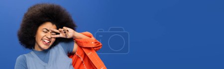 Cheerful african american woman in stylish outfit gesturing on blue background, banner 