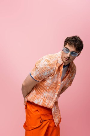 Young brunette man in sunglasses and shirt looking away isolated on pink 