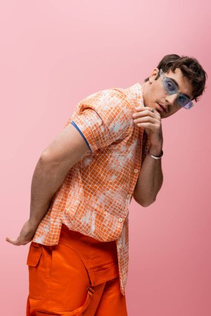 Stylish model in orange overalls and blue sunglasses looking away isolated on pink 