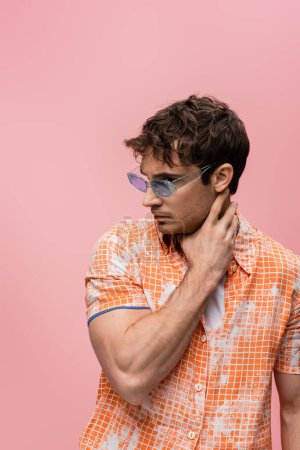 Trendy man in blue sunglasses touching neck isolated on pink 