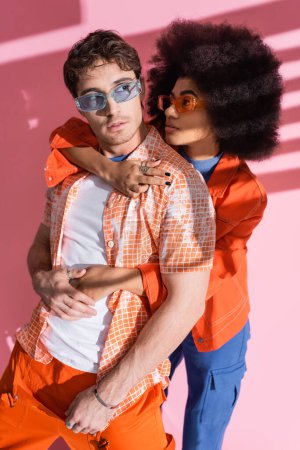 Trendy african american woman hugging man in orange overalls on pink background with shadow 