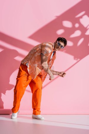 Trendy man in blue sunglasses looking at camera on pink background with shadow 