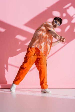 Full length of stylish man in overall and sunglasses standing on pink background with shadow 