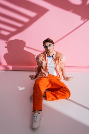 Stylish model in overall and sunglasses sitting on pink background with shadow 