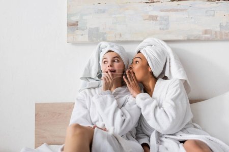 mujer afroamericana en túnica blanca contando secreto a un amigo sorprendido en la cama en casa