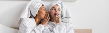 mujer afroamericana en bata de felpa y toalla contando chismes a un amigo sorprendido en el dormitorio, pancarta