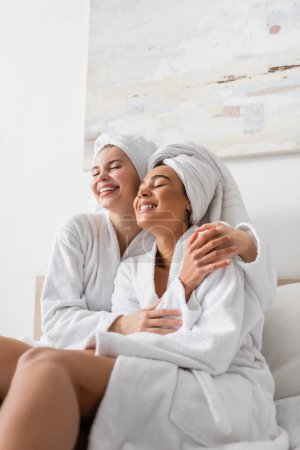 young and happy interracial women in white terry robes and towels embracing with closed eyes in bedroom