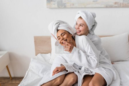 mujer afroamericana emocionada en albornoz blanco y toalla riendo con los ojos cerrados cerca de amigo feliz en la cama