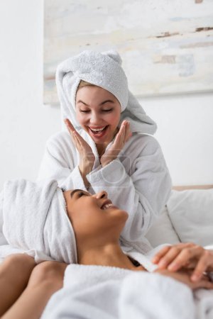 excited woman in white soft bathrobe and towel showing wow gesture near smiling african american friend in bedroom