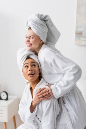 alegre joven mujer en blanco albornoz y toalla abrazando afro-americano amigo en dormitorio