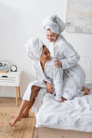 happy woman in white robe and towel embracing african american friend in bedroom