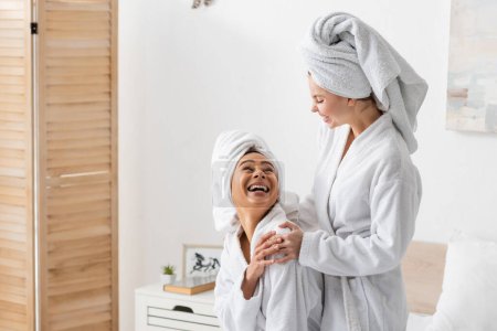 laughing african american woman in white towel and bathrobe looking at smiling friend in bedroom