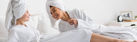 cheerful african american woman in white soft bathrobe and towel talking to young friend in bedroom, banner