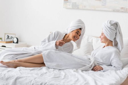 cheerful african american woman talking to pretty friend lying on bed in white terry bathrobe and towel 