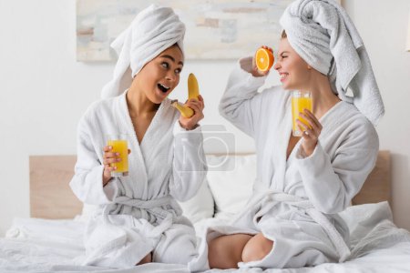 excited multiethnic women with fresh fruits and orange juice having fun while sitting on bed