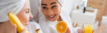 Téléchargez les photos : Joyeuse femme afro-américaine tenant de l'orange fraîche et du jus près d'un ami flou à la banane mûre, bannière - en image libre de droit