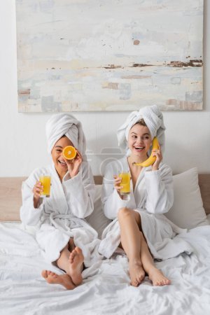 full length of cheerful interracial women with fruits and orange juice having fun while sitting on bed