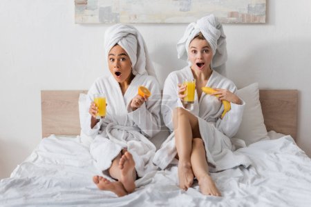 in voller Länge erstaunte interrassische Frauen in weißen Bademänteln und Handtüchern mit Früchten und Orangensaft, während sie auf dem Bett in die Kamera schauen 