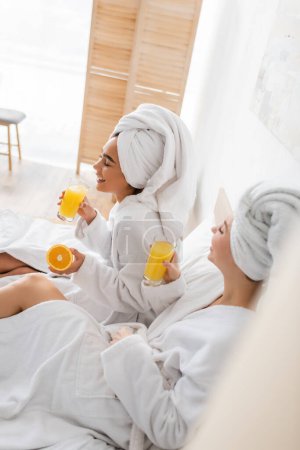 high angle view of happy interracial women in terry bathrobes and towels holding fresh orange juice on blurred foreground