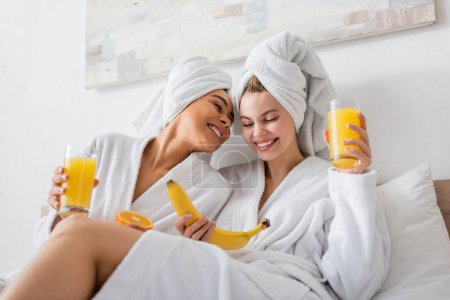 young and happy multiethnic women in white bathrobes and towels holding fruits and fresh orange juice in bedroom