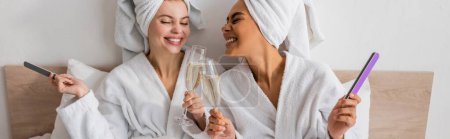 Foto de Pleased multiethnic women in terry bathrobes and towels holding nail files and clinking champagne glasses in bedroom, banner - Imagen libre de derechos