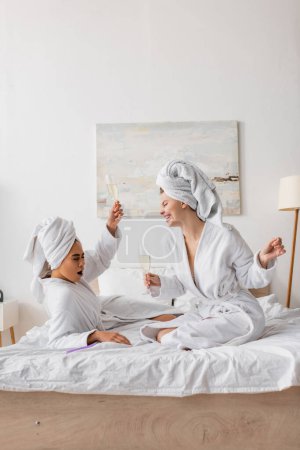 excited african american woman toasting with champagne near happy friend in white robe and towel in bedroom