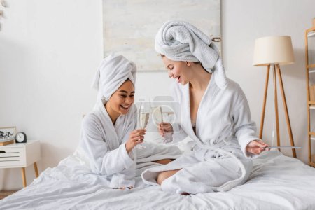 Téléchargez les photos : Gai interracial femmes en blanc doux peignoirs et serviettes clinquant verres à champagne sur le lit - en image libre de droit