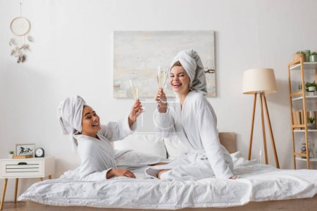 femmes multiethniques gaies en serviettes et peignoirs blancs regardant la caméra et griller avec du champagne dans la chambre moderne