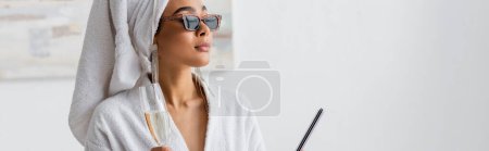 african american woman in white robe and stylish sunglasses holding champagne and nail file while looking away at home, banner