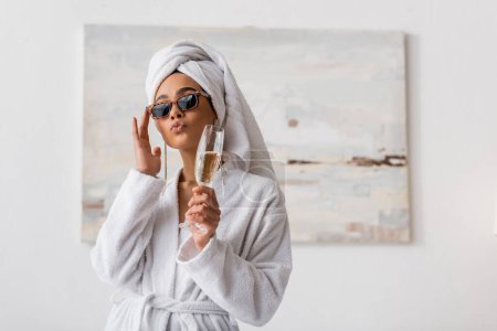 Afroamerikanerin in Frottee-Bademantel und Handtuch, die trendige Sonnenbrille anpasst, während sie Lippen schmollt und Champagner im Schlafzimmer hält