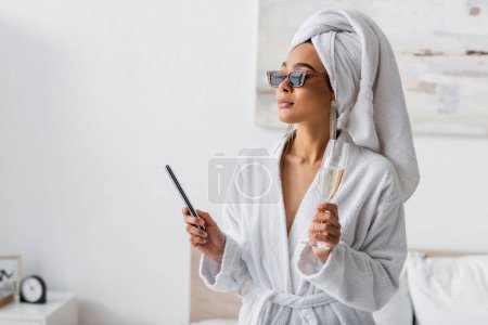 young african american woman in soft terry bathrobe and sunglasses looking away while holding nail fail and champagne in bedroom