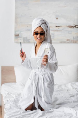 cheerful african american woman in white bathrobe and sunglasses posing with nail file and champagne glass in bedroom