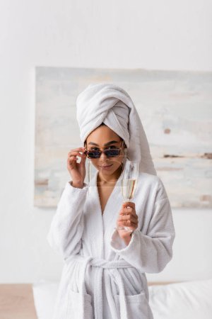 junge afrikanisch-amerikanische Frau in weißem Bademantel und Handtuch mit Champagner in der Hand und Blick in die Kamera über Sonnenbrille im Schlafzimmer