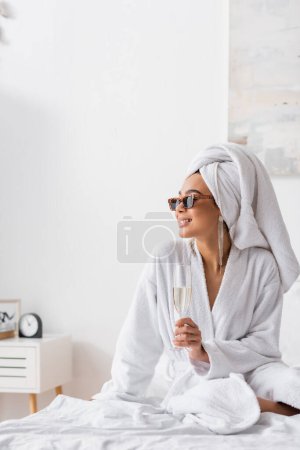 femme afro-américaine souriante en peignoir éponge et lunettes de soleil à la mode tenant champagne et regardant loin dans la chambre