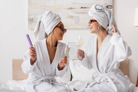 happy interracial women in terry robes and stylish sunglasses holding champagne and looking at each other in bedroom