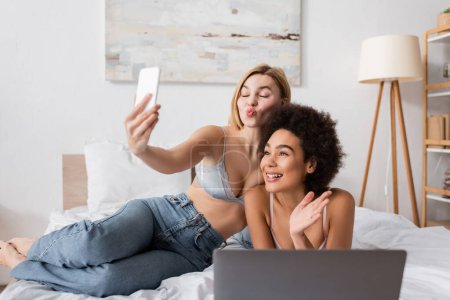 blonde woman pouting lips while taking selfie with smiling african american friend waving hand in bedroom