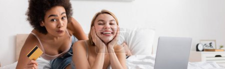 Téléchargez les photos : African american woman with credit card grimacing near happy friend and laptop in bedroom, banner - en image libre de droit