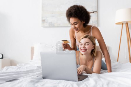 Téléchargez les photos : Heureuse femme afro-américaine tenant carte de crédit près ami souriant en utilisant un ordinateur portable sur le lit - en image libre de droit