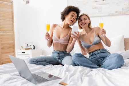 staunende und fröhliche multiethnische Frauen mit Sektgläsern mit Cocktails in der Nähe eines verschwommenen Laptops auf dem Bett