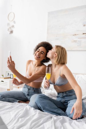 blonde Frau mit Cocktail im Champagnerglas küsst lächelnden afrikanisch-amerikanischen Freund Selfie auf dem Bett zu Hause