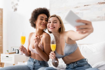happy interracial women in lingerie and jeans holding champagne glasses with cocktails and taking selfie on blurred smartphone