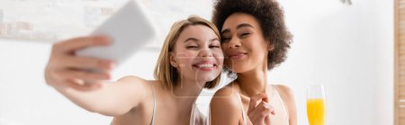 happy blonde woman taking selfie with african american friend holding champagne glass with orange juice, banner