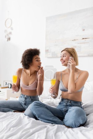 erstaunt afrikanisch-amerikanische Frau mit Cocktail schaut blonde Freundin an, die auf Handy spricht