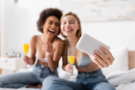 Selektiver Fokus des Smartphones in der Nähe verschwommener interrassischer Frauen mit Cocktails in Sektgläsern, die Selfies im Schlafzimmer machen