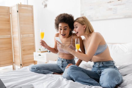 verblüffte interrassische Frauen in Dessous und Jeans, die Cocktails halten und im Schlafzimmer auf das Smartphone schauen