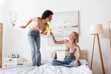 joyful interracial women in jeans and bras clinking champagne glasses with cocktails in bedroom