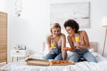 glückliche interrassische Frauen halten Champagnergläser mit Cocktails in der Hand, während sie zu Hause leckere Pizza im Bett betrachten