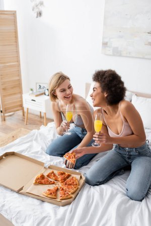 alegres mujeres interracial en sostenes y jeans sentados con cócteles en copas de champán en la cama en casa